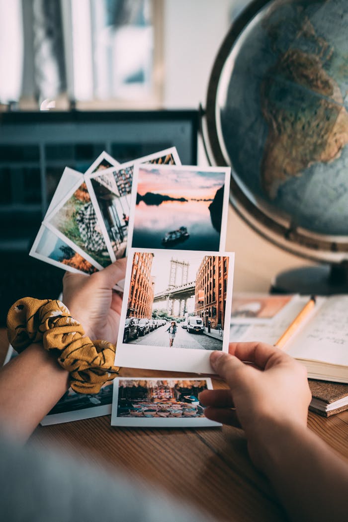 Hand holding polaroid pictures with travel theme near a globe, indoors.